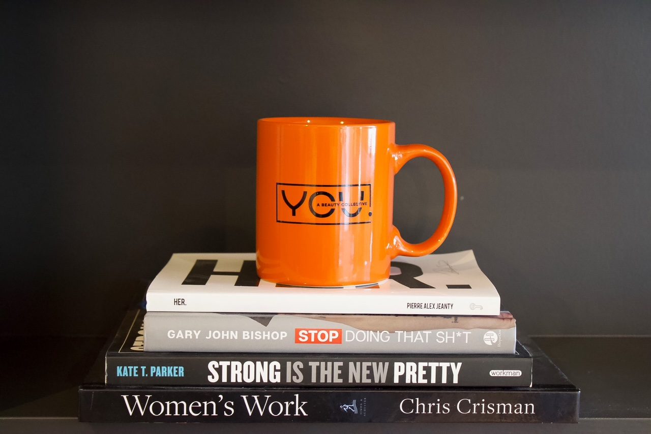 An orange mug with the YOU. MedSpa logo, placed atop stacked books, with the title "Strong is the New Pretty, Women's Work, Stop Doing that SH*T, and HER."